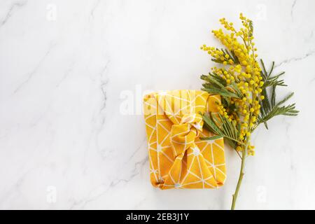 Vista dall'alto del regalo confezionato in tessuto di lino in stile furoshiki eco con fiore mimosa giallo. Concetto di celebrazione del giorno della donna o della madre. Foto Stock