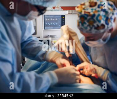 Concentratevi sul monitor della frequenza cardiaca e sul team medico che esegue un intervento di chirurgia del pancia in sala operatoria. Chirurgo in plastica e assistente che esegue interventi chirurgici sull'addome del paziente. Concetto di apparecchiatura medica. Foto Stock