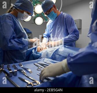 Team medico in tute chirurgiche che eseguono interventi di chirurgia estetica in sala operatoria. Medici che fanno chirurgia estetica mentre assistente in piedi da tavolo chirurgico con strumenti medici. Concetto di chirurgia plastica. Foto Stock