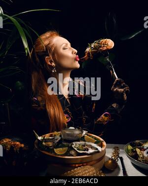 Una ricca donna dai capelli rossi sta mangiando nel ristorante di pesce che tiene un gamberetto forcato o langoutine e sta andando baciarlo. Tavolo da pranzo con cibo di mare e un Foto Stock