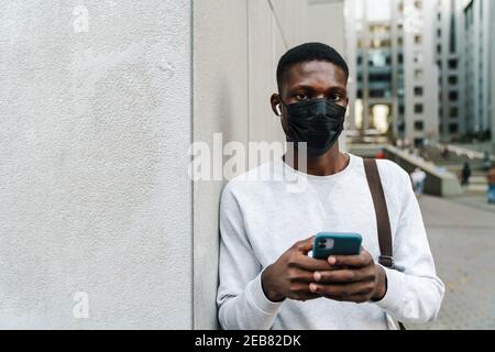 Giovane afroamericano uomo in maschera di faccia usando gli auricolari e. telefono cellulare all'aperto Foto Stock