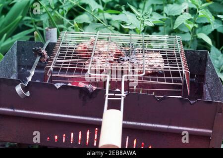 grigliate di carne in barbecue. cestello grigliato con manico in legno e spiedino piatto in acciaio su griglia a carbone. sullo sfondo di erba verde spessa Foto Stock