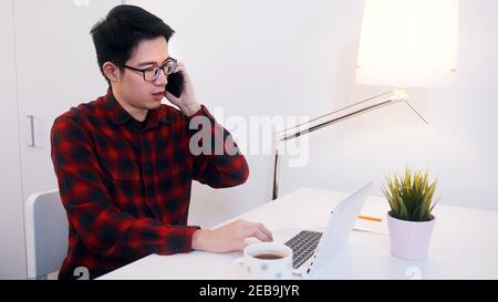 Giovane uomo d'affari asiatico che lavora sulla scrivania a casa e che ha conversazione telefonica. Foto di alta qualità Foto Stock