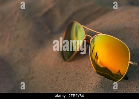 Occhiali da sole riflettenti da vicino sulla spiaggia di sabbia con il tramonto e la riflessione della gente. Occhiali da sole a specchio sulla spiaggia di sabbia in estate sera. Occhiali da sole su sa Foto Stock