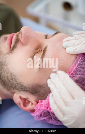 Ritratto di giovane uomo sulla procedura di pulizia del viso da parte della cosmetologa donna in clinica di bellezza. Beutician medico sta pulendo il viso del paziente con tamponi di cotone. Foto Stock