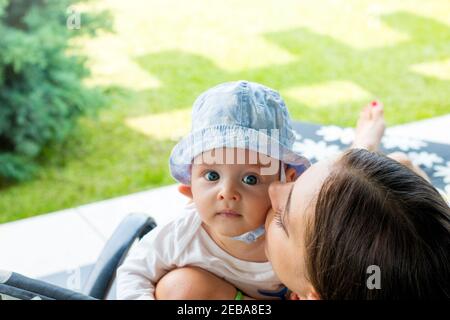 Mamma allegra abbracciando, coccolandosi e guancia baciando bambino all'aperto, ritratto di bel bambino dagli occhi blu che riposa nelle braccia amorevoli della mamma Foto Stock