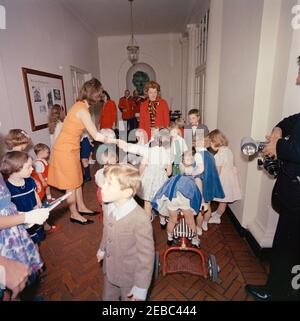 Festa di compleanno per Caroline Kennedy e John F. Kennedy, Jr. First Lady Jacqueline Kennedy (a sinistra) consegna un regalo avvolto a sua figlia, Caroline Kennedy (centro a destra, parzialmente nascosta), durante una festa di compleanno congiunta per Caroline e John F. Kennedy, Jr.; John, Jr. (Di nuovo alla macchina fotografica), si trova a destra di sua madre. Il fotografo della Casa Bianca, Captain Cecil Stoughton (per lo più fuori dal quadro), si trova all'estrema destra; i membri della United States Marine Band giocano sullo sfondo. East Wing Corridor, Casa Bianca, Washington, D.C. Foto Stock