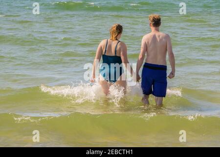 Amata coppia che va a nuotare in onde spruzzi di mare. Donna e uomo si preparano per nuotare in mare ondulato. Coppia che tiene insieme le mani andando più lontano Foto Stock