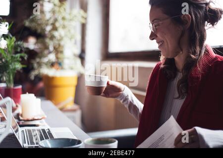 la donna sorride con una tazza di tè mentre lavora su un computer portatile in ufficio a casa Foto Stock