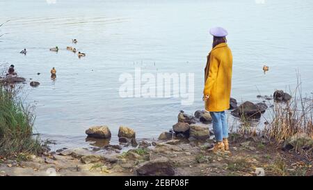 donna in elegante abito autunnale in piedi vicino al lago con selvaggio anatre nel parco Foto Stock