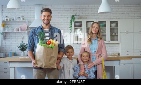 Uomo sorridente che tiene un sacchetto di carta con cibo fresco vicino alla famiglia con bambini in cucina Foto Stock