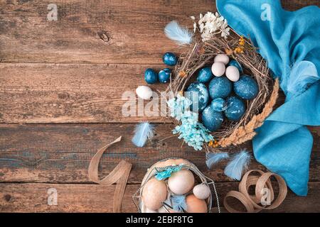 Uova di Pasqua blu-marmo in un nido di vimini con piume e fiori accanto a un tovagliolo blu e un cestino di uova pastello. Foto Stock