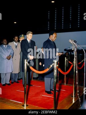 Cerimonia di arrivo per Kwame Nkrumah, presidente del Ghana, 15:43. Il presidente della Repubblica del Ghana, Osagyefo Dr. Kwame Nkrumah, fa commenti alle cerimonie di arrivo in suo onore. Da sinistra a destra: Due uomini non identificati; il presidente dei capi di Stato Uniti, il generale Lyman Lemnitzer (parzialmente nascosto); il presidente John F. Kennedy; il segretario di Stato, Dean Rusk (dietro il braccio del presidente Kennedyu2019s); il presidente Nkrumah (ai microfoni). Hangar n. 10, Terminal del Servizio militare di trasporto aereo (MATS), Aeroporto Nazionale di Washington, Washington, D.C. Foto Stock