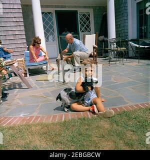 Viaggio a Hyannis Port u2013 Squaw Island, 1963-08-14. John F. Kennedy, Jr., gioca con il suo cane, Shannon, a casa Brambletyde su Squaw Island, Hyannis Port, Massachusetts. Il presidente John F. Kennedy parla al telefono all'estremità sinistra della cornice; First Lady Jacqueline Kennedy e l'assistente presidenziale, Dave Powers, giocano con il cane della famiglia Kennedy, Wolf, in background. Foto Stock