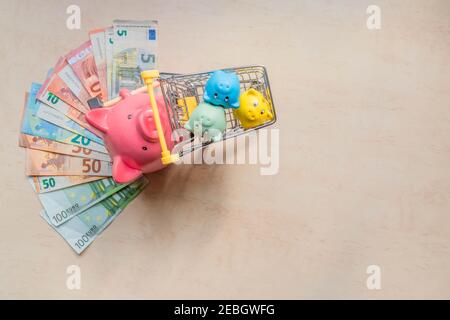 Una grande banca di maiale che rotola tre piccole banche di maiale nel carrello di shopping su sfondo di legno con banconote in euro. Grandi banche di suino e suinetti, banconote in euro e s. Foto Stock