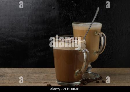 Due tazze in vetro con cappuccino e cioccolato ghiacciato su un tavolo di legno con fondo a trama nera. Messa a fuoco selettiva. Foto Stock