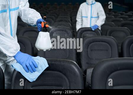 Due lavoratori in indumenti protettivi sedie di pulizia in sala cinema con disinfettanti. Concetto di disinfezione. Foto Stock