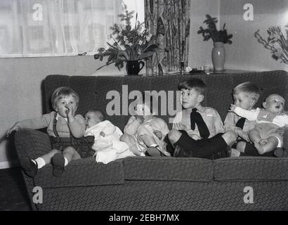 Anni '50 storico, in una stanza di fronte, su un divano, sei bambini giovani seduti insieme in una fila, due ragazzini, due bambine e due bambini, forse per una foto di famiglia, Inghilterra, Regno Unito. Far sedere insieme sei bambini fianco a fianco è stato un successo. Foto Stock