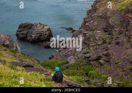 Ragazzo in felpa con cappuccio seduto sulla costa rocciosa dell'Atlantico il giorno nuvoloso, St. Bride's, Terranova Foto Stock