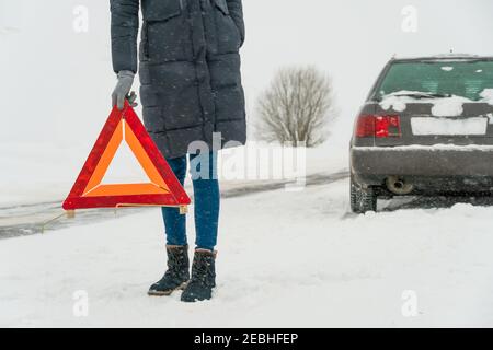 Donna mette in primo piano un cartello di avvertimento triangolare su una strada in una giornata invernale innevata. Concetto di guasto dell'auto Foto Stock