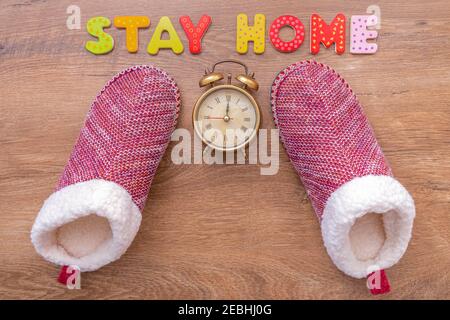 Le parole RIMANGONO CASA di lettere di legno, sveglia e pantofole su sfondo di legno. Text STAY HOME con accessori per la casa su pavimento in legno. Vista dall'alto, copia Foto Stock