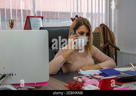 La donna di affari che indossa la maschera protettiva del viso parla dal telefono a. la sua scrivania Foto Stock