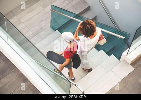 Alla moda di madre e figlio camminando sulle scale in un moderno contesto della città. Ragazzo con lo skateboard va giù la scaletta con mamma vista dall'alto di persone passeggiate in Foto Stock