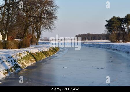 Un canale ghiacciato nei Paesi Bassi accanto ai campi innevati sotto un cielo blu d'inverno Foto Stock