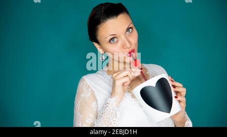 Donna caucasica che guarda la macchina fotografica mentre beve su una paglia da una grande tazza con un cuore. Foto Stock