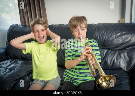 Due bambini seduti su un divano. Uno sta suonando una tromba e l'altro bambino infastidito sta coprendo le orecchie con le mani Foto Stock