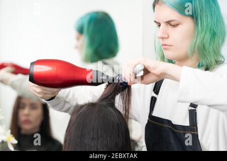 Professionale parrucchiere con capelli verdi asciuga i capelli al cliente con asciugacapelli rosso e pettine blu in salone di bellezza. Foto Stock