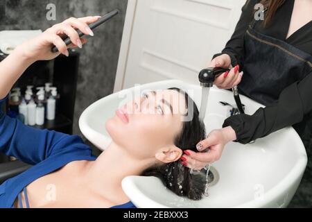 Il cliente guarda il telefono mentre il parrucchiere è occupato lavando i suoi capelli al salone di bellezza. Le mani del hairstylist lavano i capelli lunghi della donna della brunetta con shampoo Foto Stock