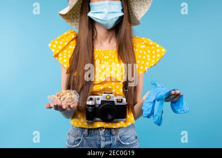 Foto ritagliata di donna brunetta con fotocamera che indossa maschera medica protettiva e cappello estivo, tenendo guanti medici blu e conchiglie marine. Pandemia Foto Stock