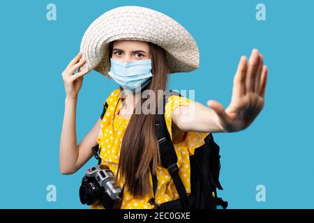 Donna turistica con fotocamera e zaino che indossa cappello estivo e maschera medica per prevenire il coronavirus in resort, mostrando stop, gesto di avvertimento Foto Stock