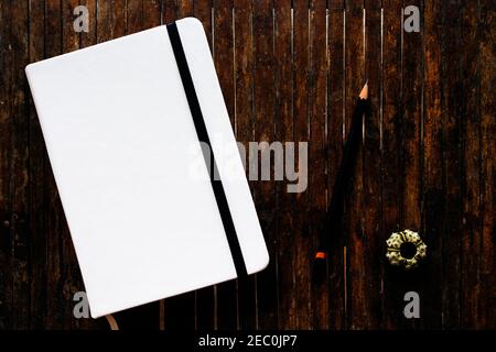 Copertina bianca abbozzetto con matita nera su tavola rustica in legno piatto posa foto. Notebook chiuso con copertina vuota e foto in posizione piana. Blocco note sul piano del tavolo Foto Stock