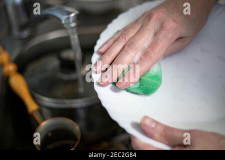 una donna lava i piatti con una spugna di schiuma, tiene un piatto nelle mani sopra il lavandino, l'acqua scorre dal rubinetto Foto Stock