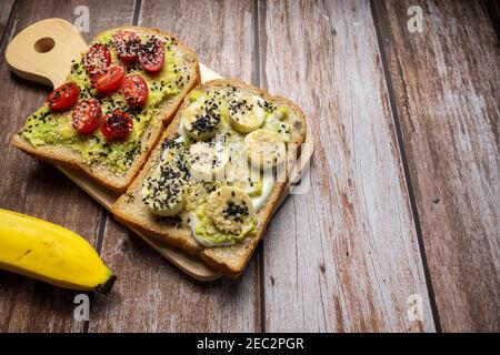 Colazione sana con toast avocado, pomodoro, banana, yogurt con pane sandwich a grani interi. Concetto di alimentazione sana, dieta, vegeta vegana Foto Stock