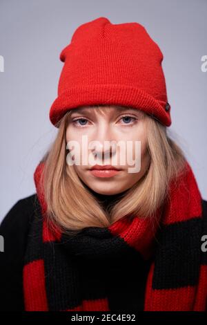 Ragazza bionda con capelli lunghi che esprime emozione neutra. Bella modello femminile in berretto rosso e sciarpa guardando dritto in macchina fotografica Foto Stock