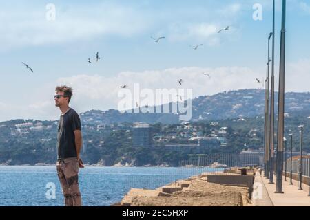 Giovane uomo in piedi sul molo di cemento di mare in porto e guardando avanti. Gabbiani e gabbiani nel cielo. Uomo su sfondo stagcape. Linea mediterranea, Palamo Foto Stock