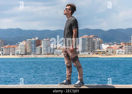 Giovane uomo in piedi sul molo del mare e guardando avanti, esprimendo felicità e libertà. Uomo su sfondo stagcape. Migrazione e nuovo concetto iniziale. Foto Stock