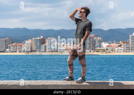 Giovane uomo in piedi sul molo del mare e guardando avanti, esprimendo felicità e libertà. Uomo su sfondo stagcape. Migrazione e nuovo concetto iniziale. Foto Stock