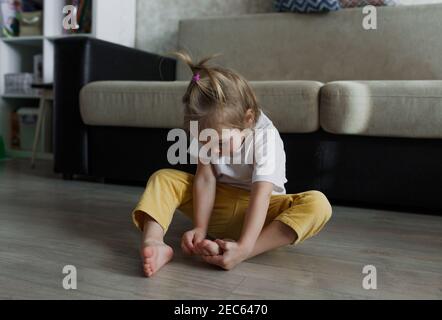 Una bambina in una T-shirt bianca e pantaloni gialli si siede sul pavimento della casa ed esamina attentamente le sue gambe. Foto Stock