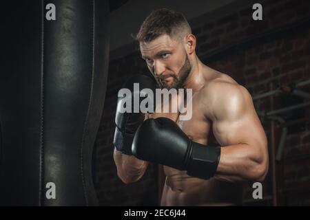 combattente con aggressività batte la borsa da pugilato in guanti neri in palestra. boxer allenarsi duro, sudore andare e pelle scintillante, spazio copia Foto Stock