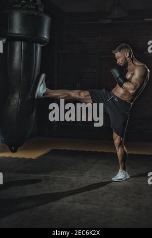 pugile in guanti neri di pugilato batte dalla borsa punzonatrice della gamba in palestra. boxer allenarsi duro e con aggressività, sudore andare e pelle scintillante, copia sp Foto Stock