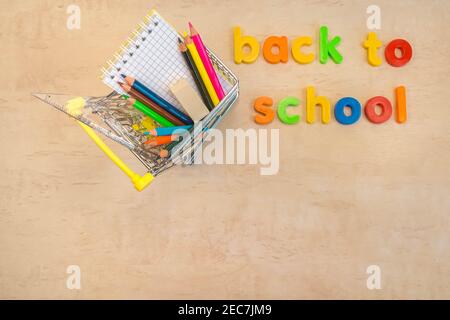 Concetto di ritorno a scuola. Posa piatta di forniture scolastiche nel carrello della spesa su sfondo di legno con testo di lettere colorate. Spazio di copia. Istruzione scolastica Foto Stock