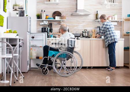 Uomo portatore di handicap in sedia a rotelle che apre il frigorifero e aiuta la moglie a preparare la colazione in cucina. Donna anziana che cucinava per il marito paralizzato, che vive con un uomo disabile con disabilità motorie Foto Stock