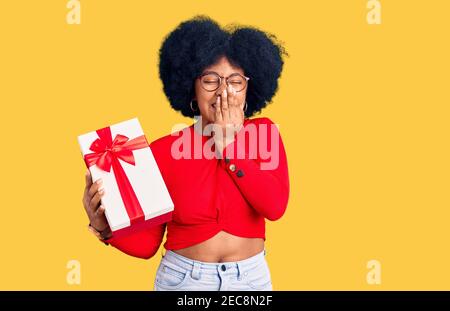 Giovane afroamericana ragazza che tiene il regalo ridendo e imbarazzato ridere che copre la bocca con le mani, pettegolezzi e concetto di scandalo Foto Stock