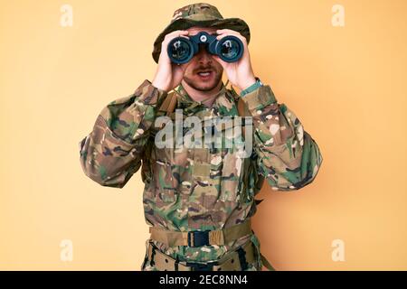 Giovane uomo caucasico che indossa uniforme armata camuffata con binocoli in faccia d'urto, che guarda scettico e sarcastico, sorpreso con la bocca aperta Foto Stock