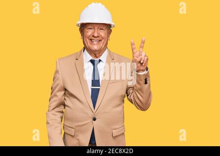 Uomo caucasico senior che indossa l'architetto hardhat sorridendo con felice faccia winking alla macchina fotografica facendo segno di vittoria. Numero due. Foto Stock