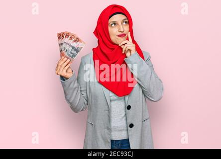 Bella donna ispanica che indossa hijab islamico tenendo in mano le banconote australiane dollari faccia seria pensare alla domanda con la mano sul mento, pensieroso Foto Stock